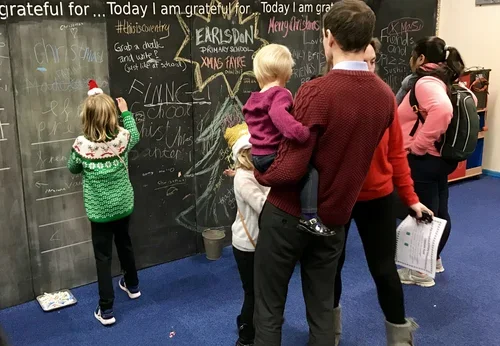 Children and a man writing on a large black chalkboard wall in a classroom, with various messages and drawings, some children wearing Christmas sweaters and backpacks.