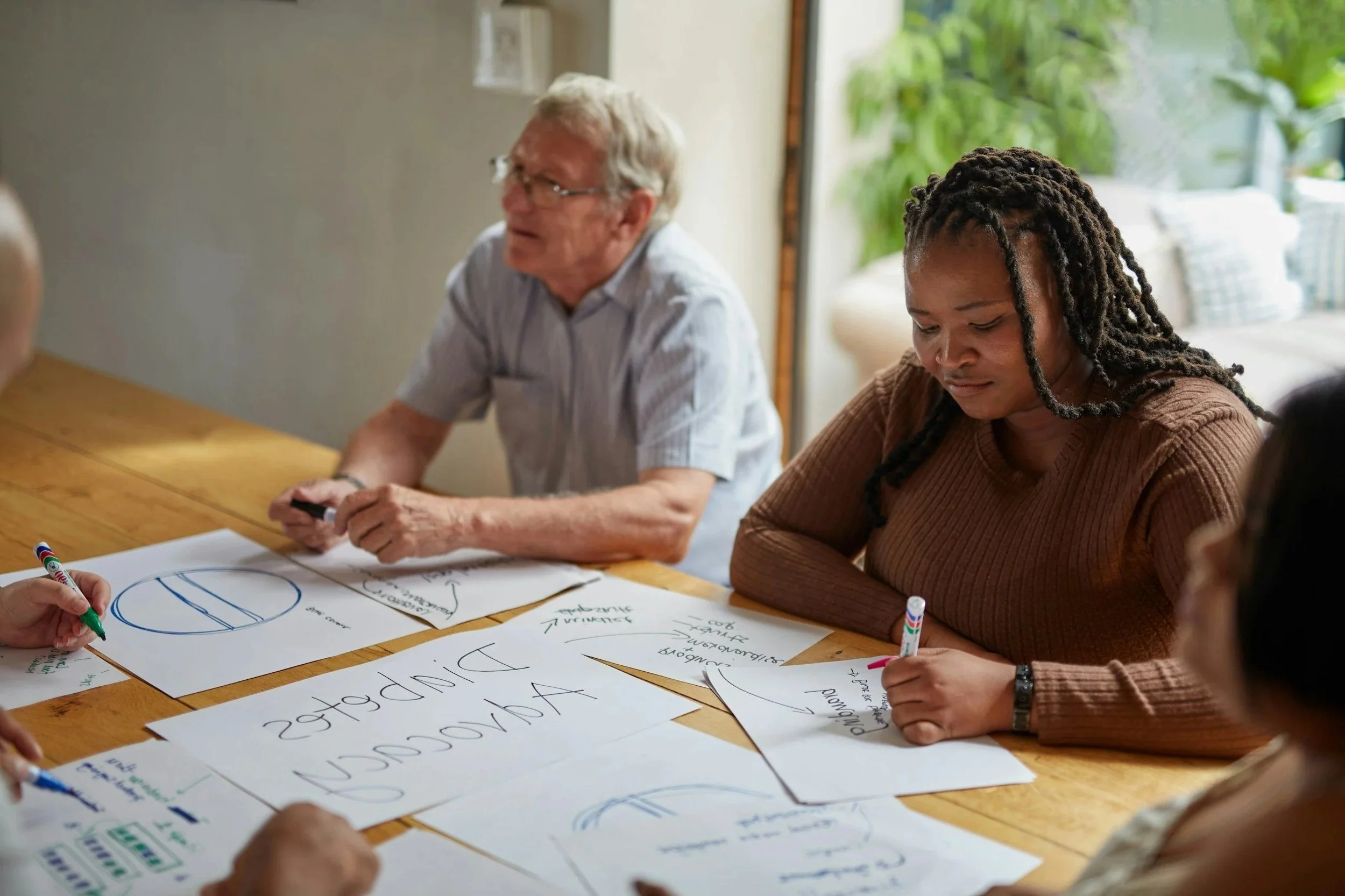 Group of people sitting around a wooden table engaged in a collaborative activity, with posters and markers on the table, in a bright room with greenery visible through a window.