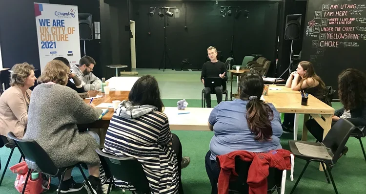A group of people sitting around tables in a room, listening to a person speaking or presenting at the front. There is a sign that reads 'WE ARE UK CITY OF CULTURE 2021' and a chalkboard with handwritten notes.