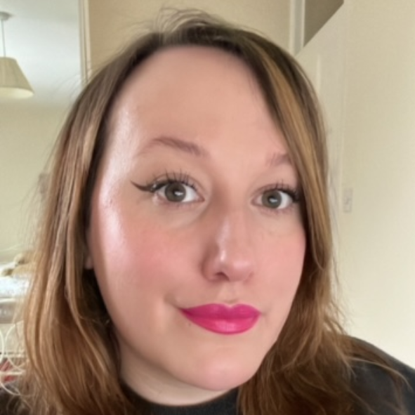 Close-up of a woman with shoulder-length light brown hair, wearing pink lipstick and black eyeliner, smiling softly indoors.