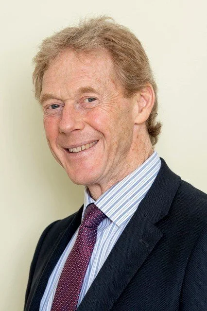 Portrait of an older man with light brown hair, smiling, wearing a dark suit, light blue shirt, and purple tie against a plain background.