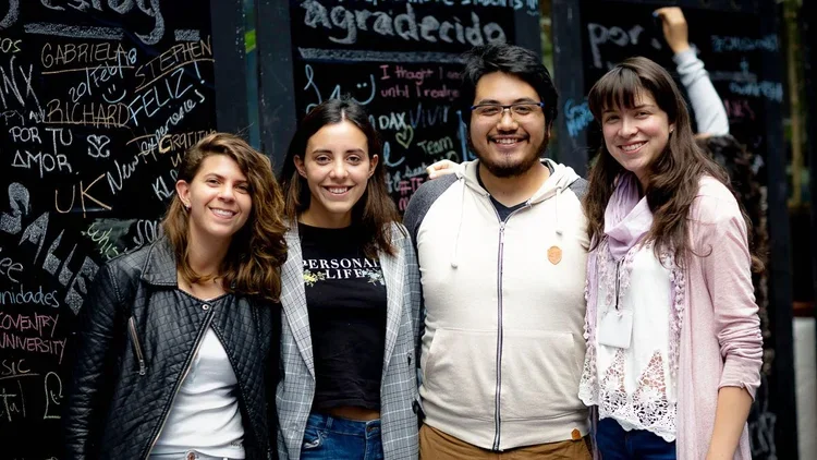 Four young people standing close together in front of a chalkboard wall with writing and signatures, smiling at the camera.