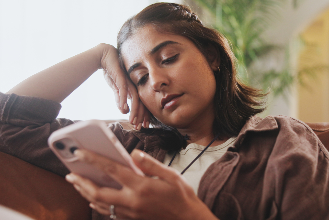 A young woman sitting on a couch, looking at her phone with a bored or tired expression, resting her head on her hand.