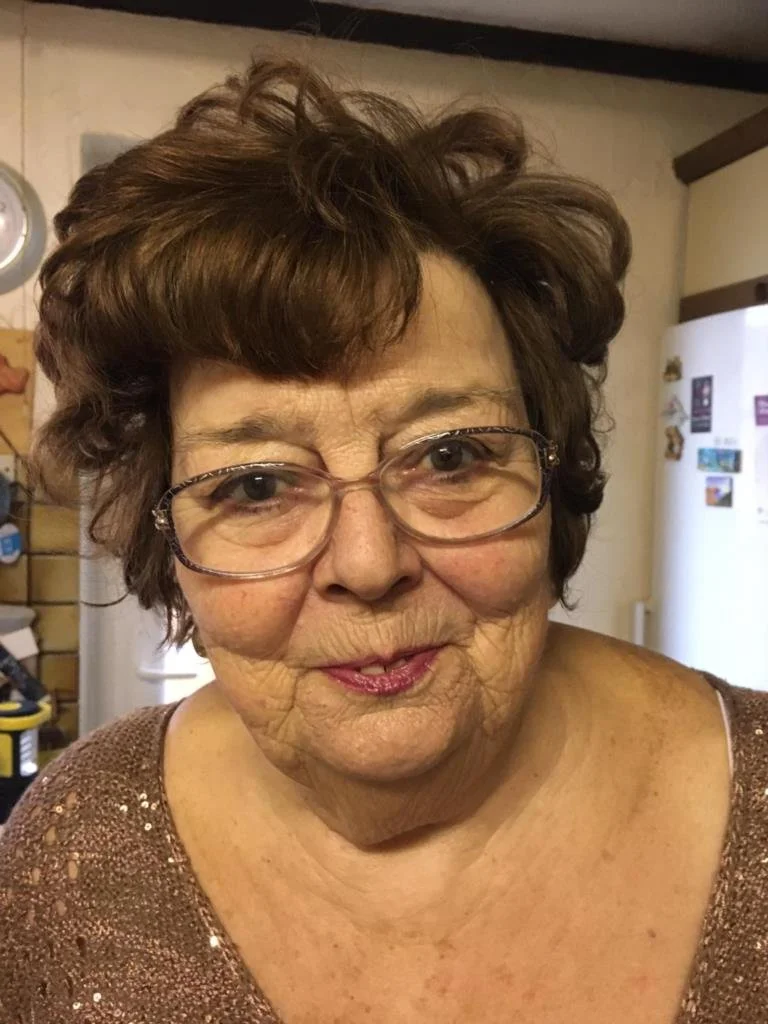 Close-up of an elderly woman with short, curly brown hair, glasses, and wearing a brown, sparkly top.