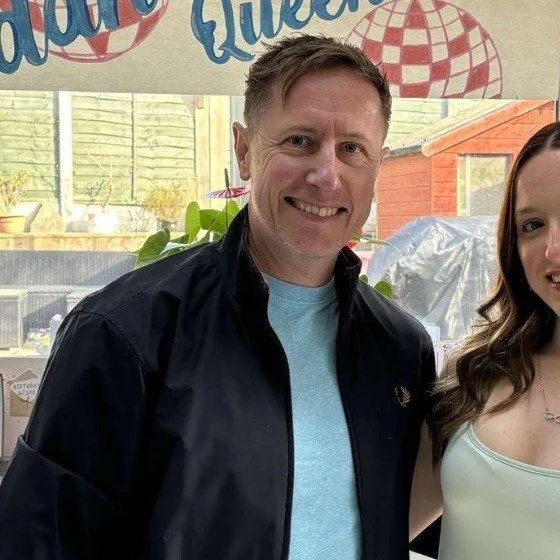 A man smiling, wearing a black jacket and a light blue shirt, standing next to a woman with long wavy hair in a white top. They are indoors.