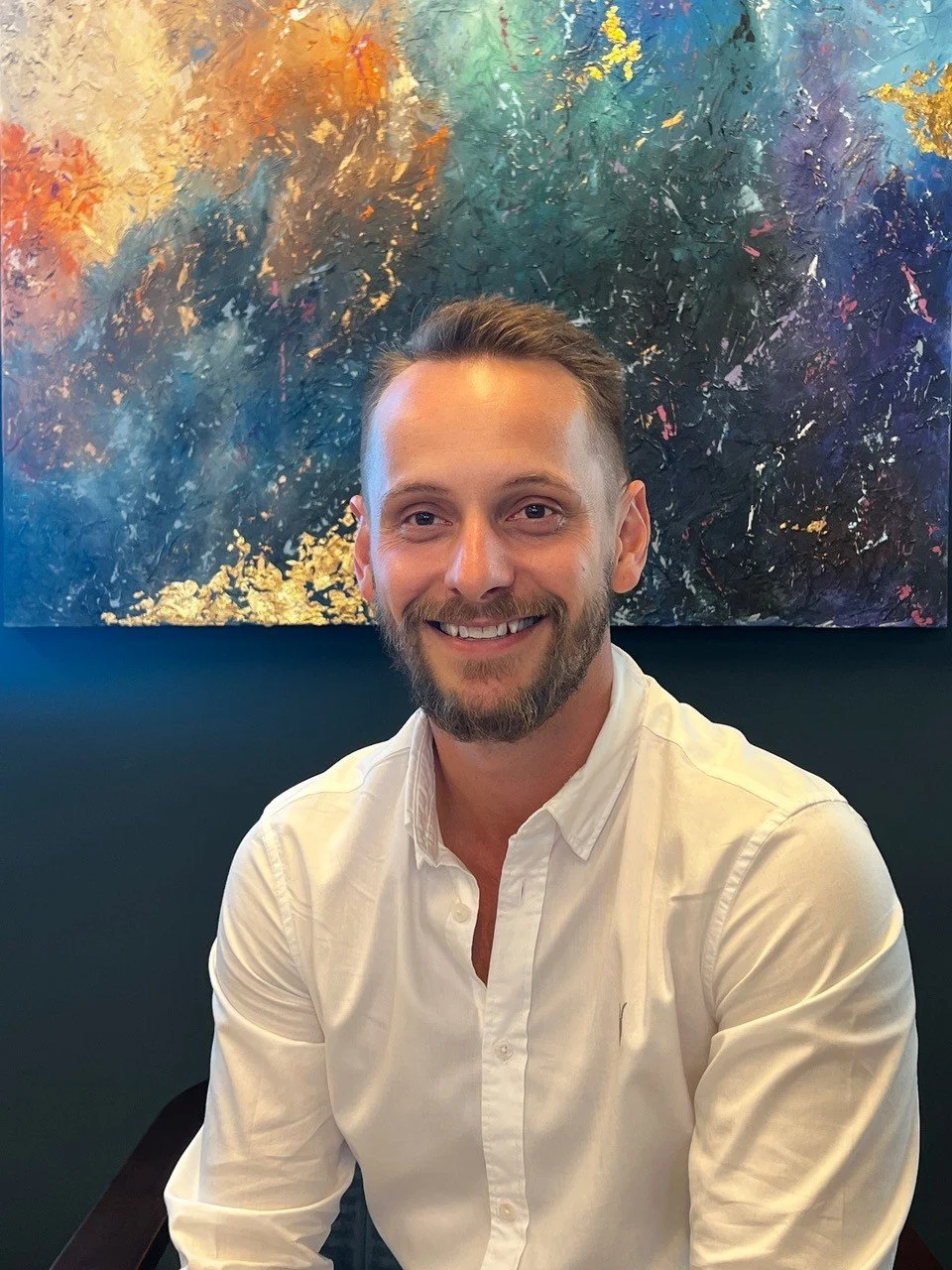 A man with short brown hair and a beard, wearing a white button-up shirt, smiling at the camera. Behind him is a colorful abstract painting with blue, orange, yellow, and purple hues.