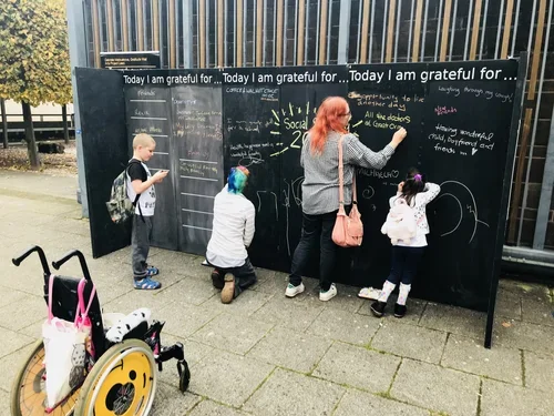 A group of children and a woman writing on a large black chalkboard outdoors, which has the phrase 'Today I am grateful for...' written at the top. A wheelchair with a pink and black bag is nearby.