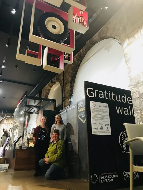 Three women standing in front of a gratitude wall inside a museum with artistic ceiling decor.