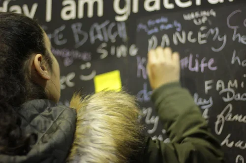 A person writing on a black chalkboard with a yellow sticky note attached, with various handwritten words in different colors, including 'gratitude' and 'dance.'