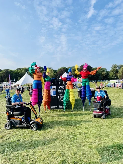 Two individuals in mobility scooters posing with colorful sea life giant puppets at an outdoor event under a blue sky.