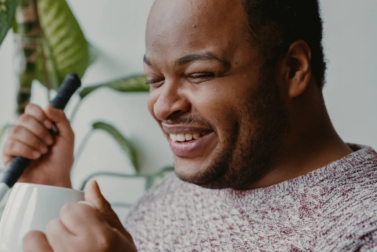A visually impaired man smiling with eyes closed, holding a mug in one hand and a guide cane in the other, with a large green plant in the background.