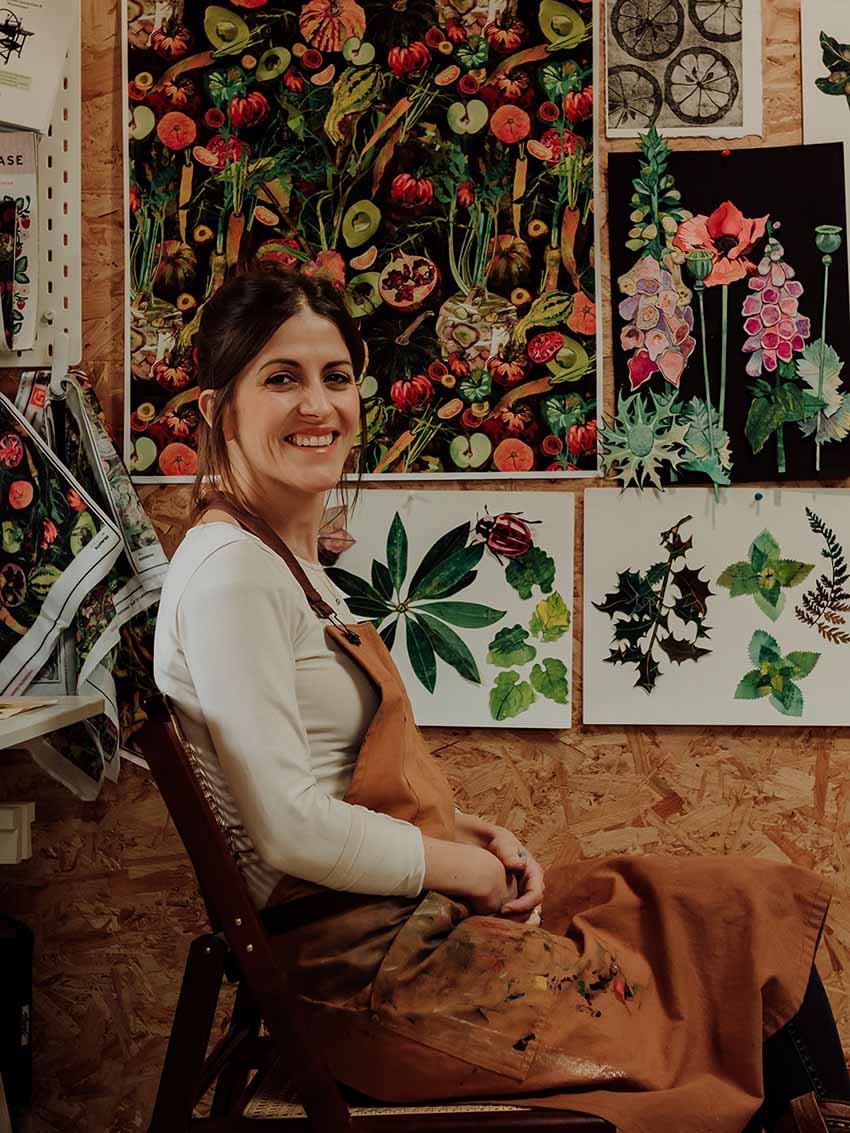 A woman wearing a paint-stained apron sitting in front of botanical artwork in an art studio.