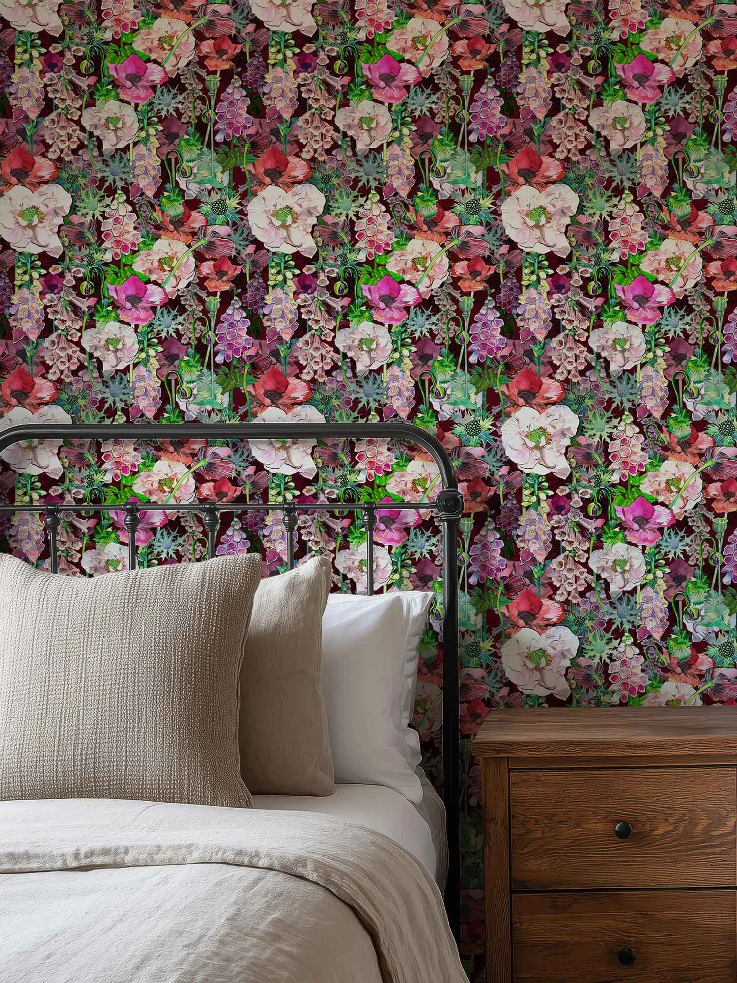 A bedroom scene with a bed covered in white linens and beige pillows, a wooden side table with drawers to the right, and a floral wallpaper with pink, purple, and green flowers in the background.