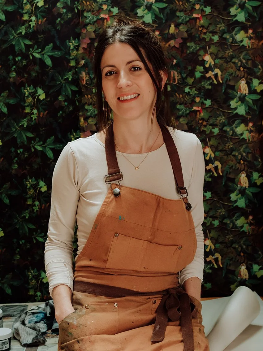 Fiona McTaggart with dark hair smiling, wearing a beige long-sleeve shirt and a brown apron, standing in front of a leafy background with small illustrations of children and animals.