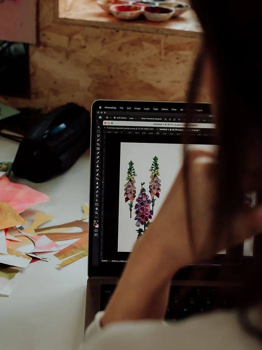 A person working on a laptop with an image of purple and pink flowers on the screen, surrounded by paper scraps on a desk, in a setting with wooden walls and small bowls of paint in the background.