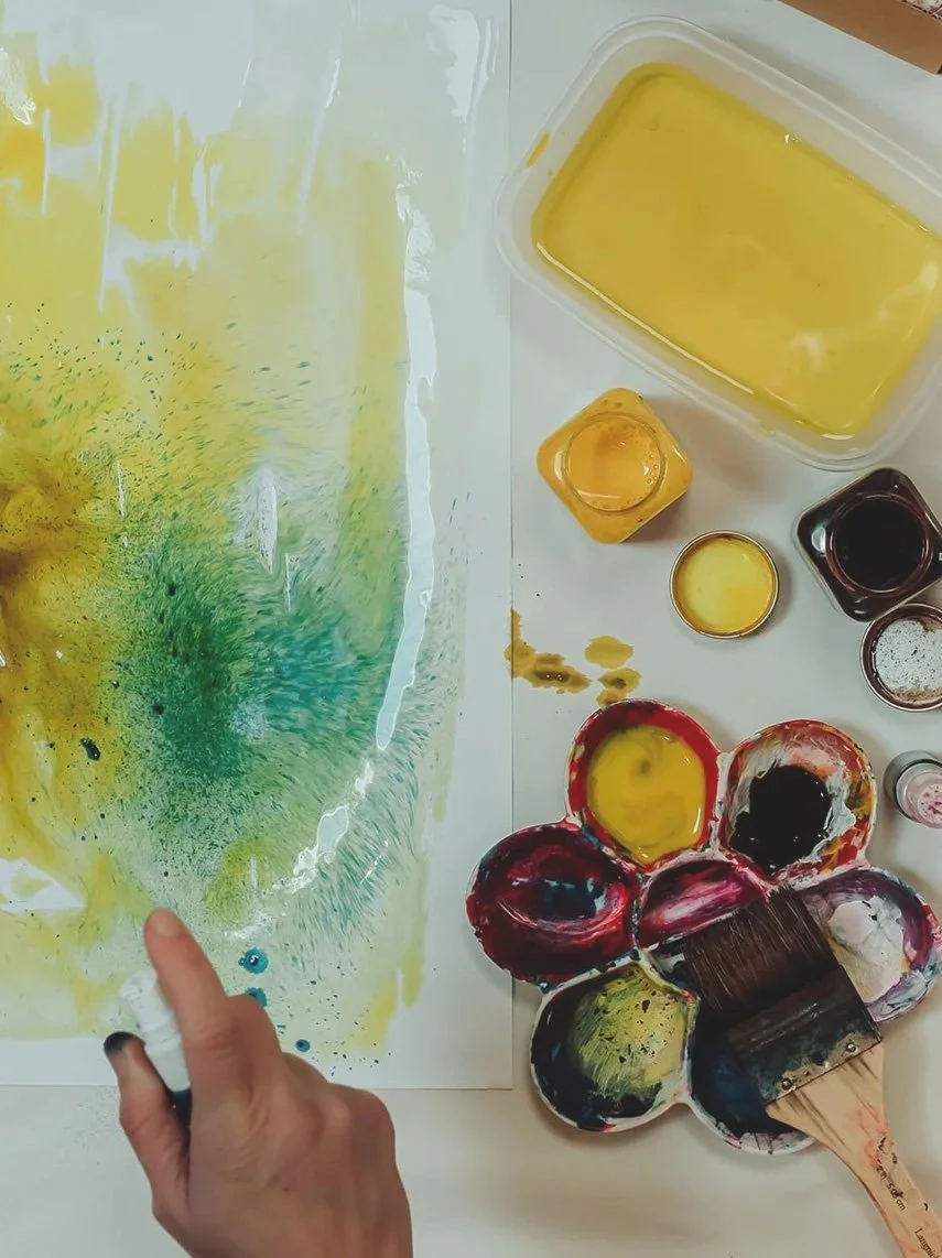 A person mixing yellow and green watercolor paints on paper with a paintbrush. Art supplies, including small containers of yellow, black, and other colored paints, are arranged on a white surface.