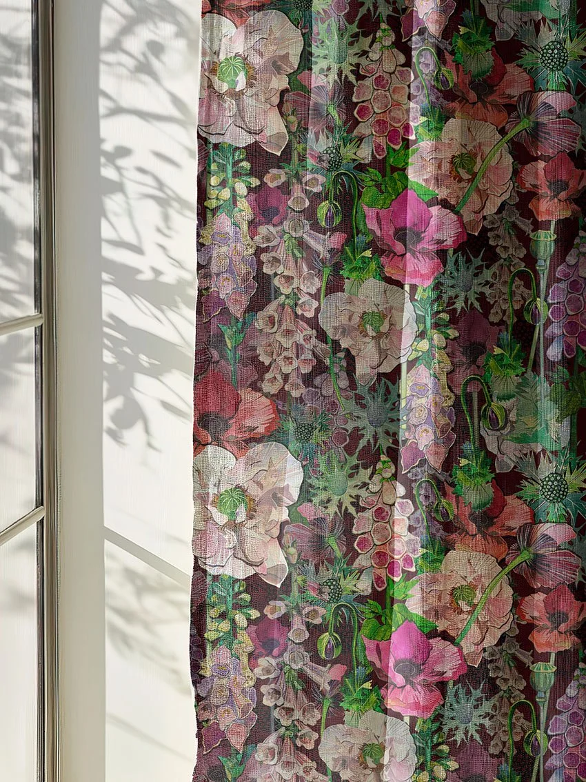 Decorative fabric curtain with a floral pattern of pink, purple, cream, and green flowers, hanging in front of a window with sunlight and shadows.