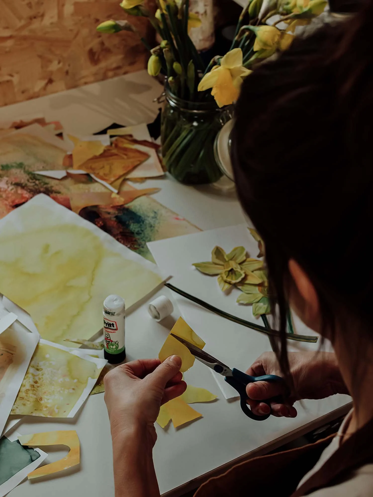 A person is cutting a yellow paper with scissors, surrounded by watercolored paper pieces and art supplies on a table, with a vase of yellow flowers in the background.