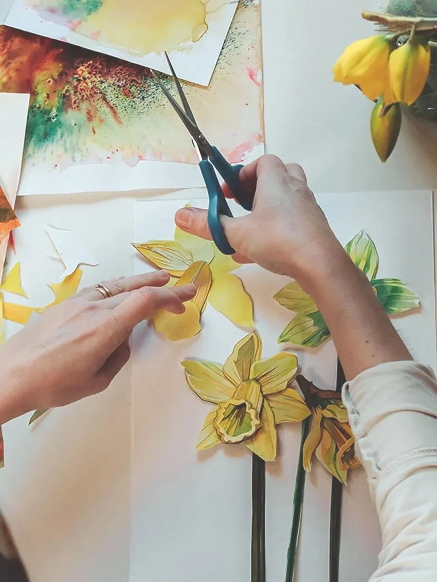 A person is cutting yellow paper flowers with scissors for a craft project, surrounded by watercolor paintings and a flower in a pot.