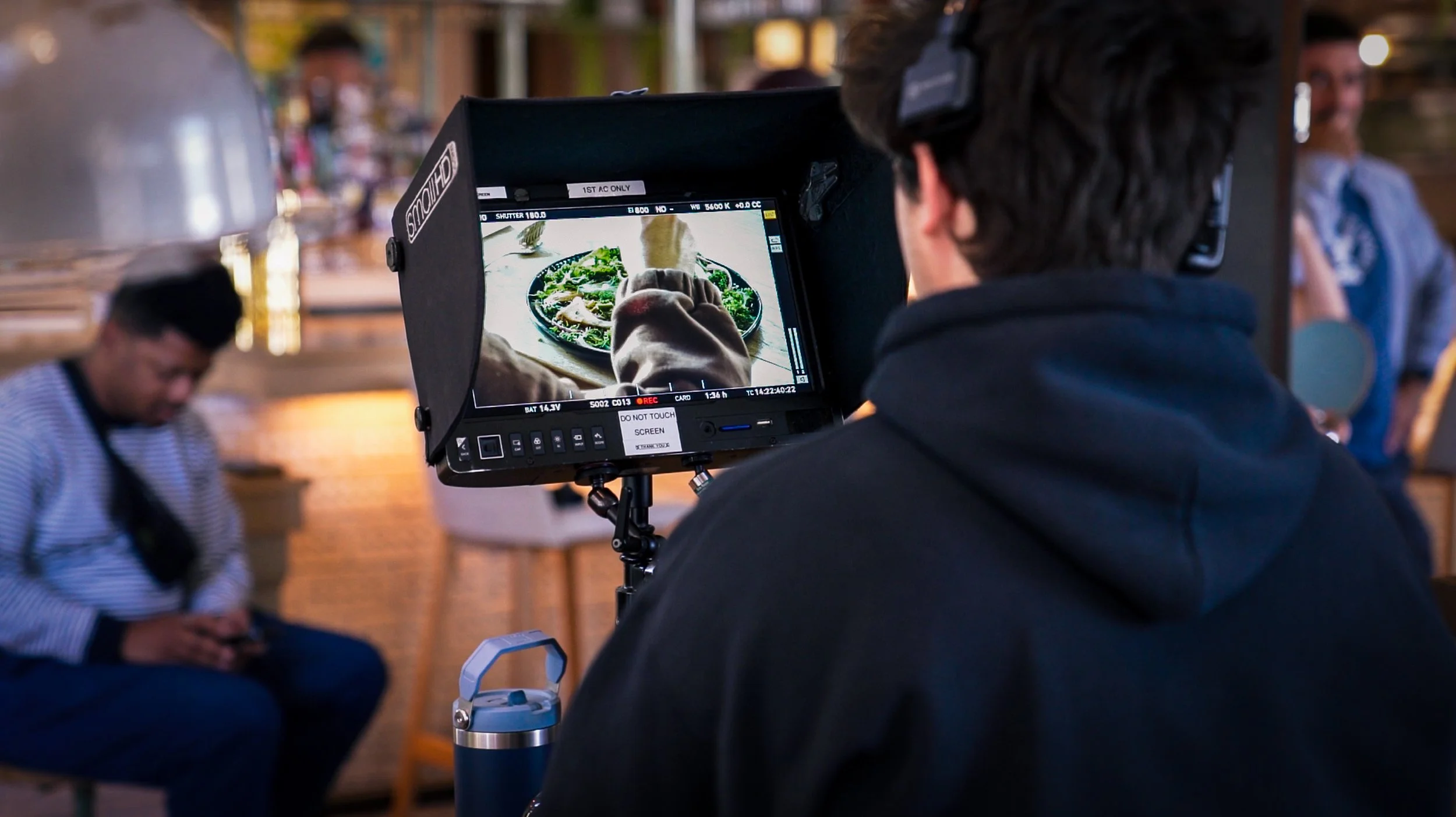 Camera operator filming a person seated at a table with a bowl of food in a restaurant or cafe setting, with people in the background.