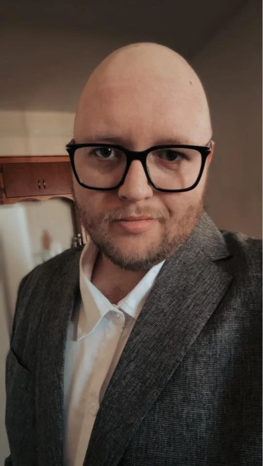 A man with a shaved head, glasses, and a beard taking a selfie indoors. He's wearing a white shirt and a gray blazer.