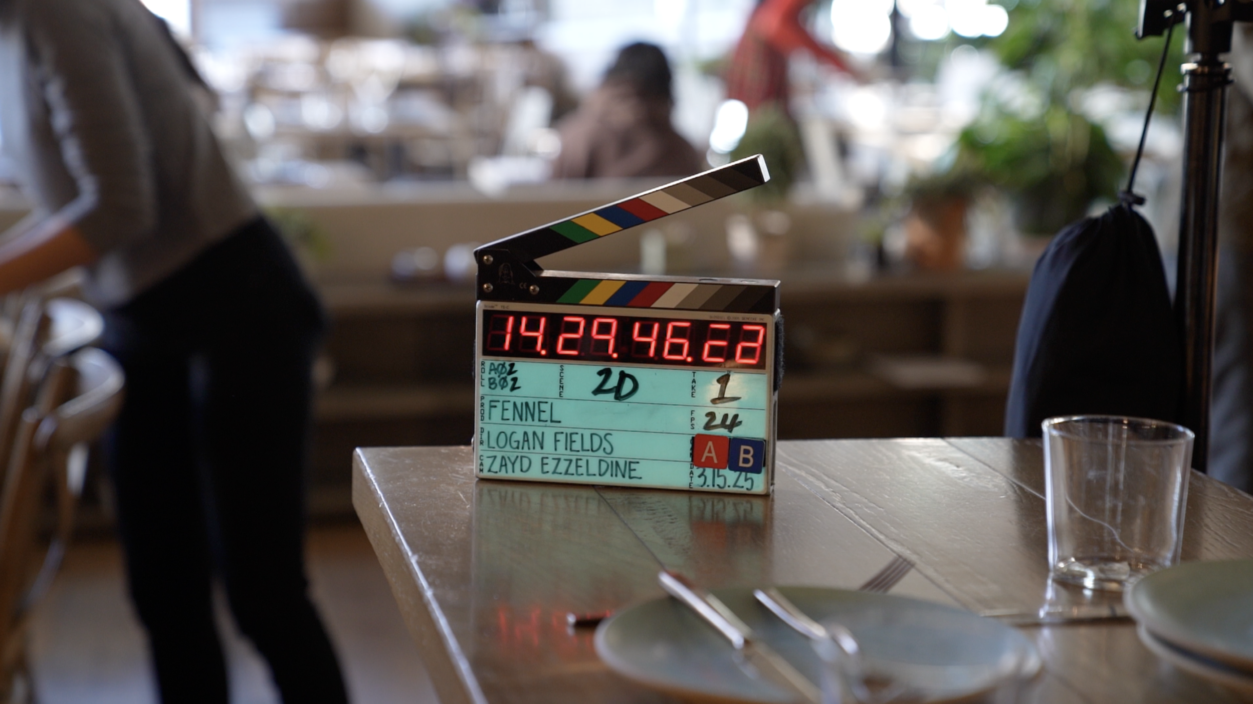 Clapperboard with digital timer and handwritten notes on a table in a restaurant or cafe setting.