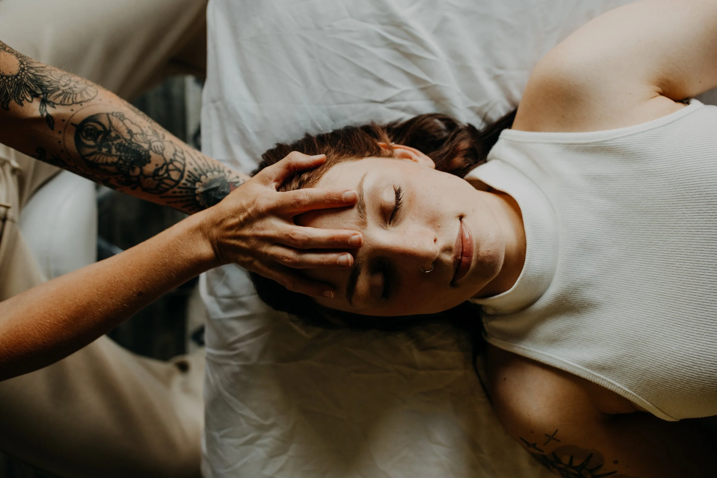 A woman with closed eyes and a nose ring lying on a bed, receiving a gentle massage on her forehead from someone with tattooed arm.