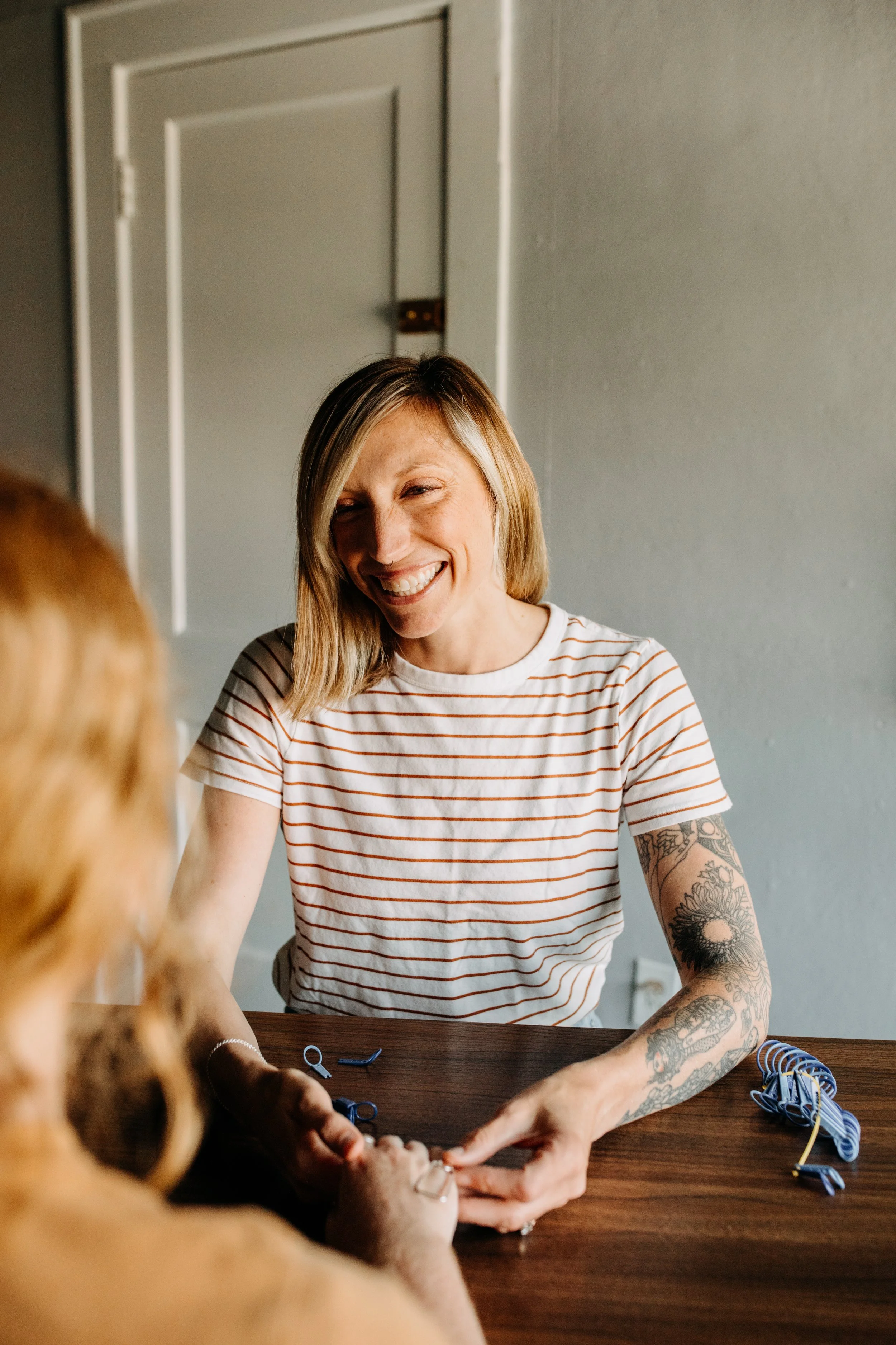 A woman with shoulder-length blond hair and tattoos on her arms is smiling and engaging in an activity with another person, whose arm is visible. They are sitting at a wooden table with some small objects and rubber bands, in a room with gray walls and a closed door in the background.