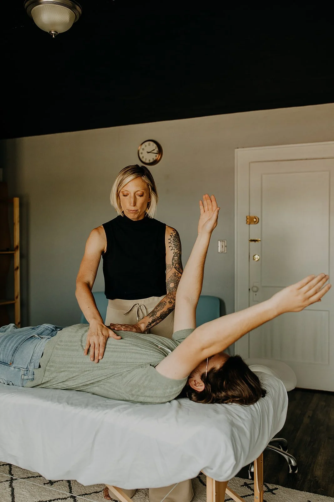 A woman giving a massage to a person lying on a massage table in a room with a clock on the wall.