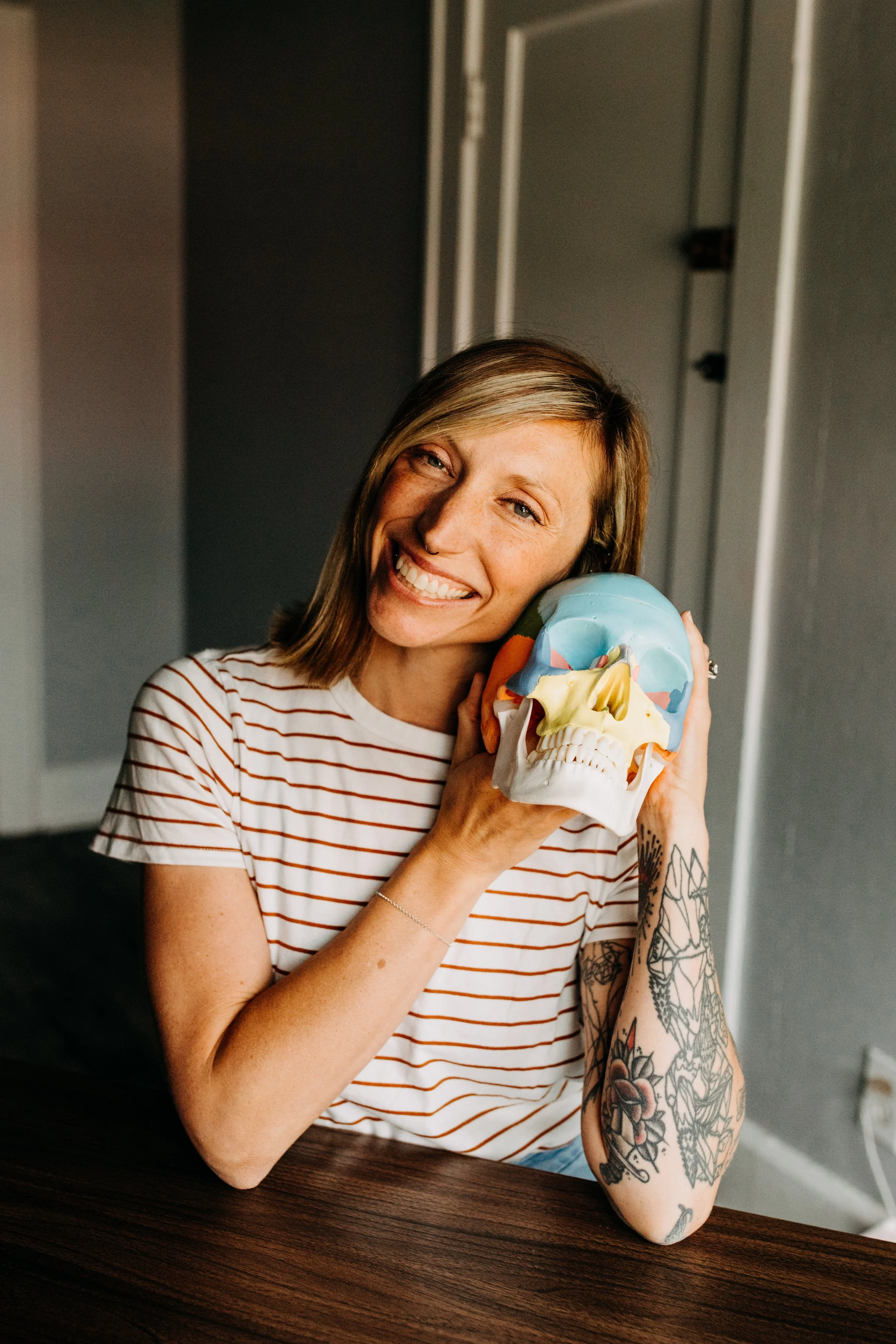 A smiling woman with tattoos on her arm is holding a colorful skull sculpture close to her face, sitting at a wooden table in a room with dark and light-colored walls.