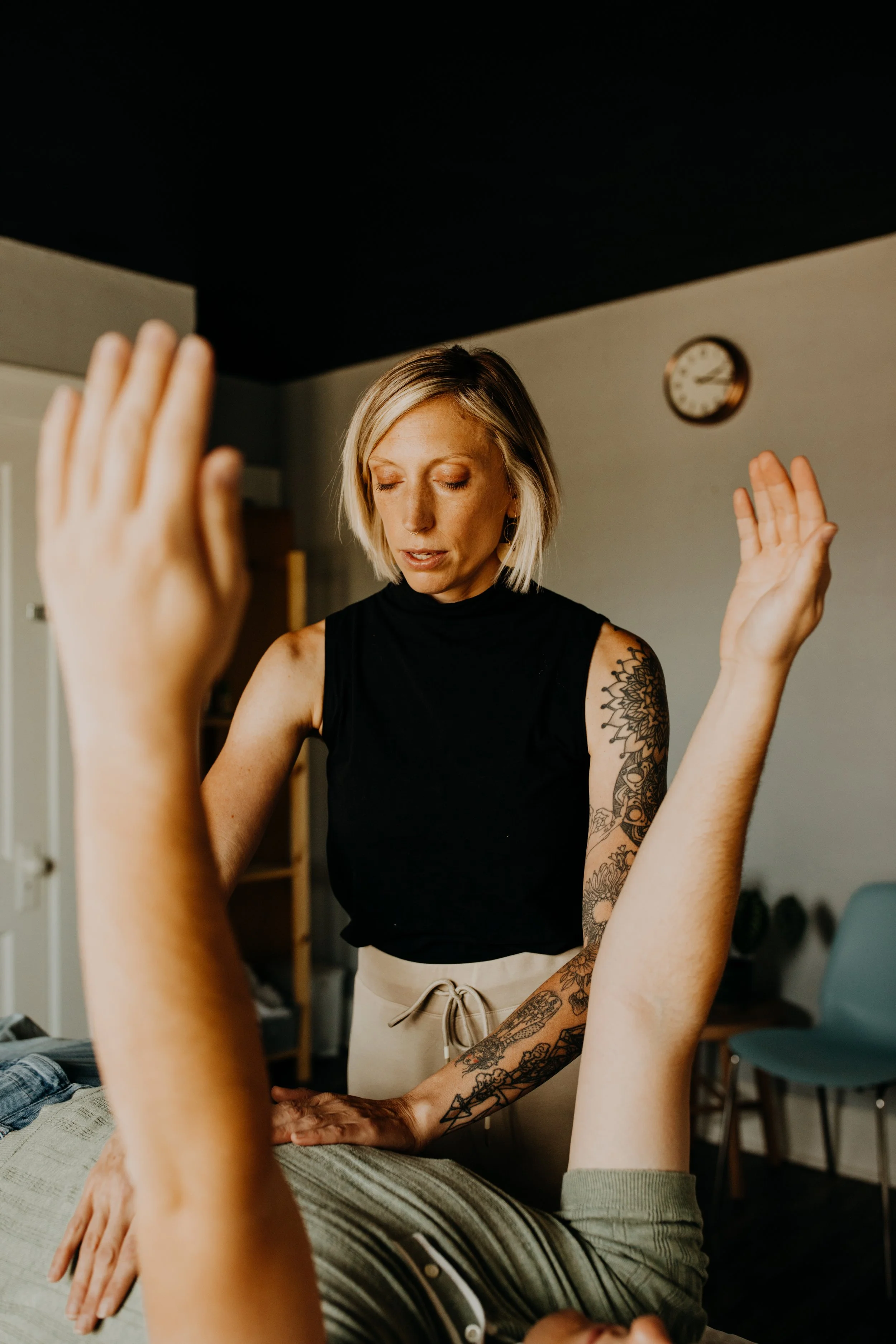 A woman with tattoos on her arm and shoulder is receiving a massage or therapy in a room with a clock on the wall.