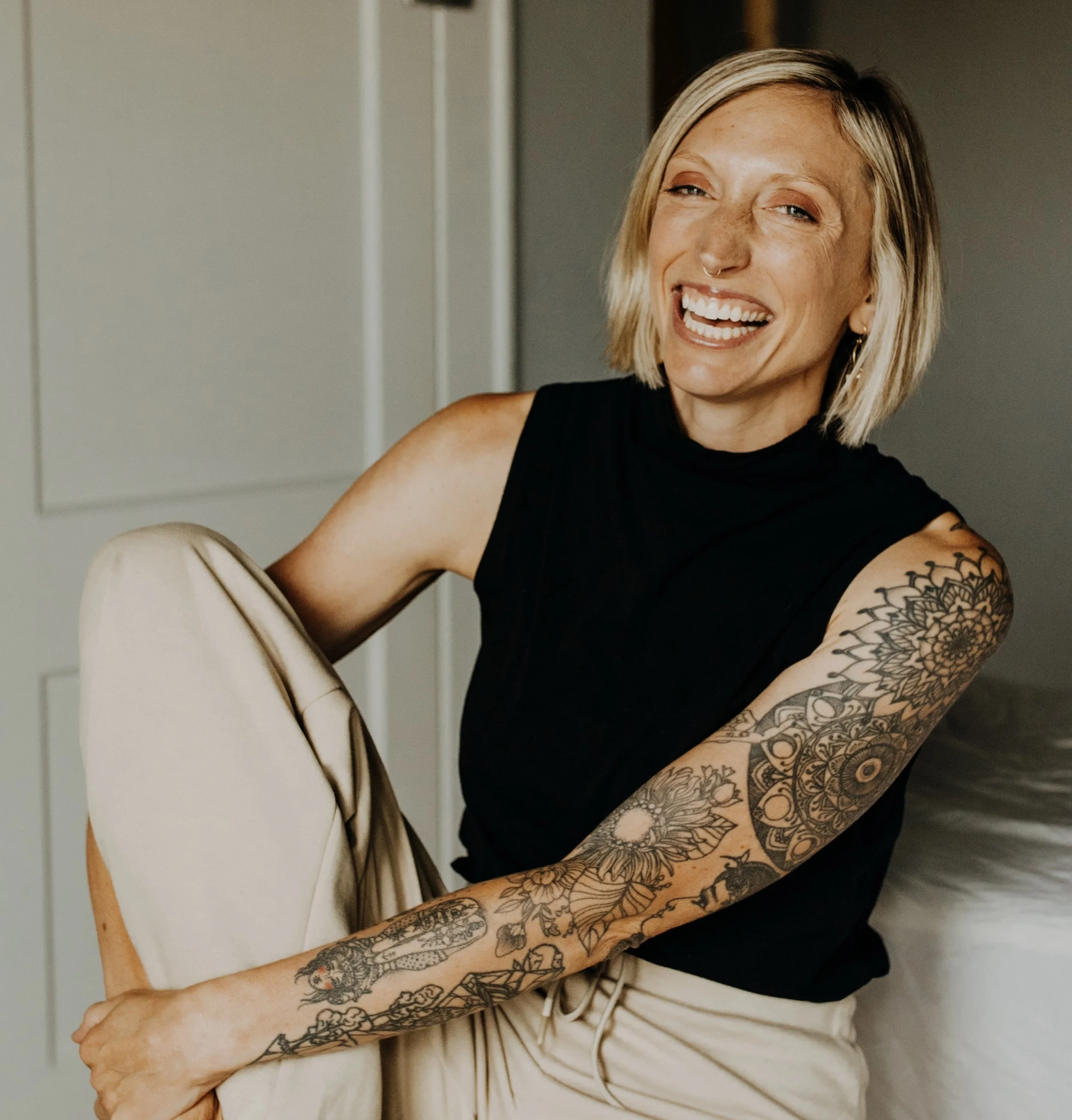 A woman with short blonde hair, wearing a black sleeveless top, smiling and sitting on a bed, showing her tattooed arm.