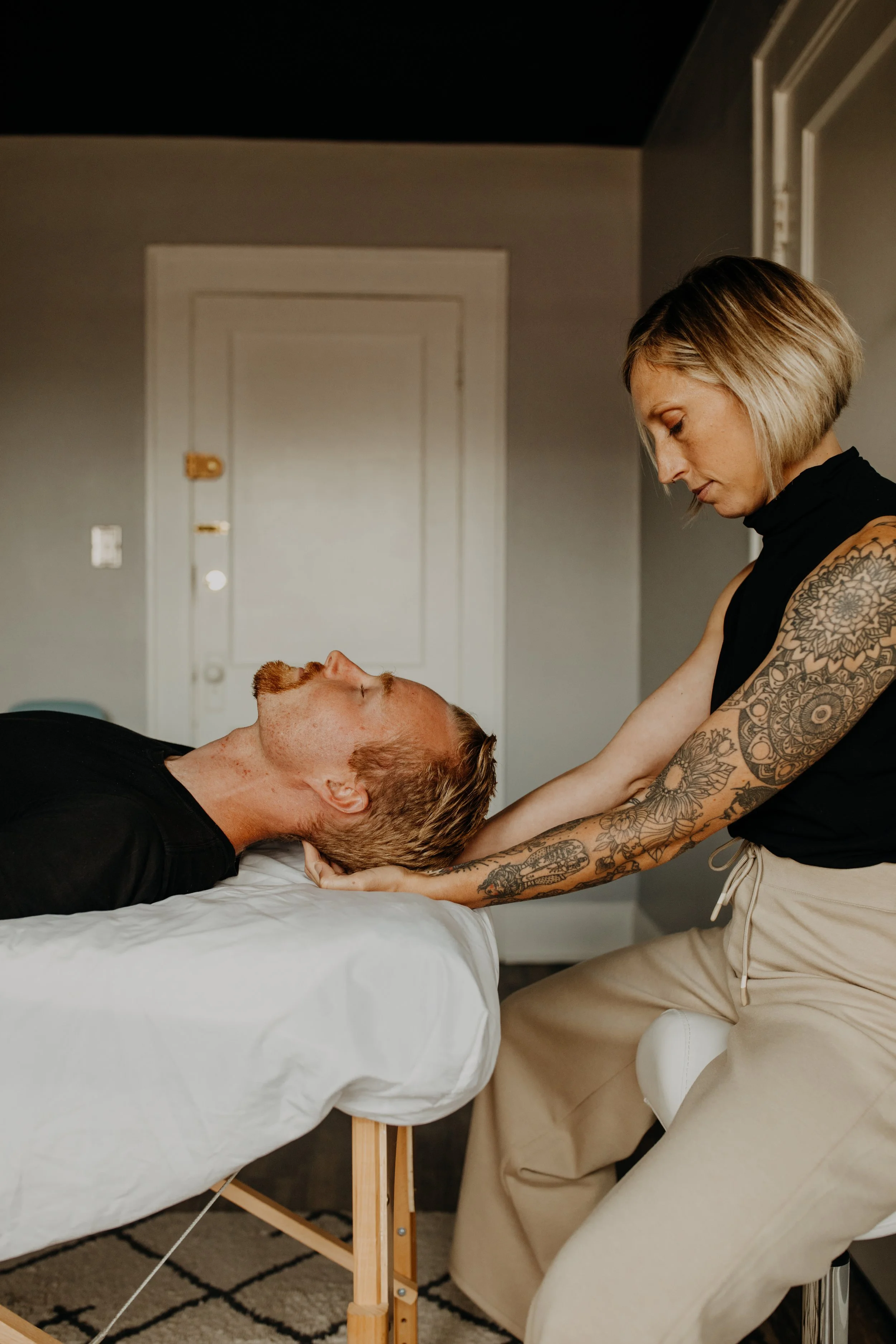 A woman with short blonde hair and tattoos on her arms is giving a massage to a man with reddish hair and a beard who is lying on a massage table with his eyes closed.