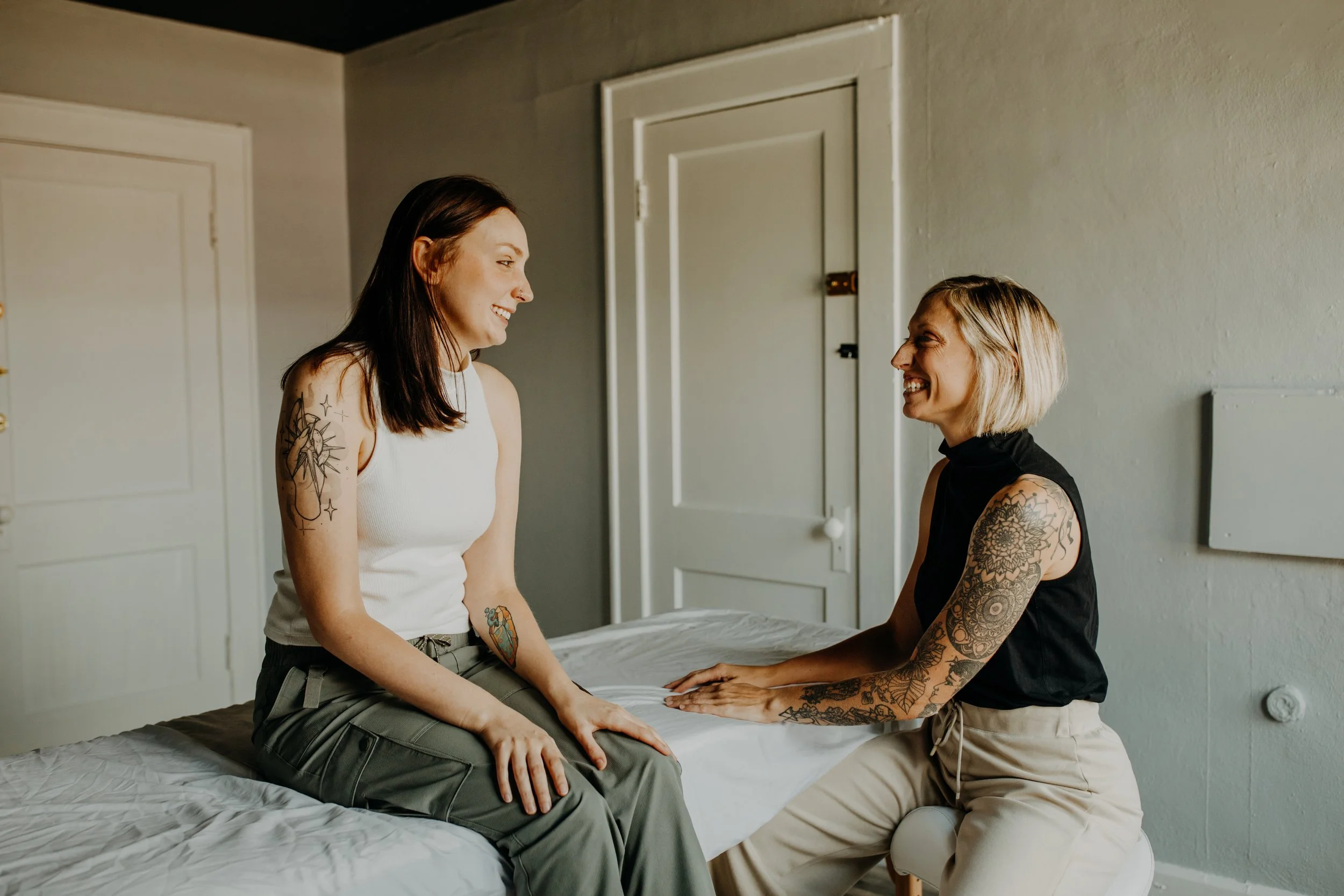 Two women smiling and talking inside a bedroom, one seated on a bed and the other on a small stool, both with visible tattoos.