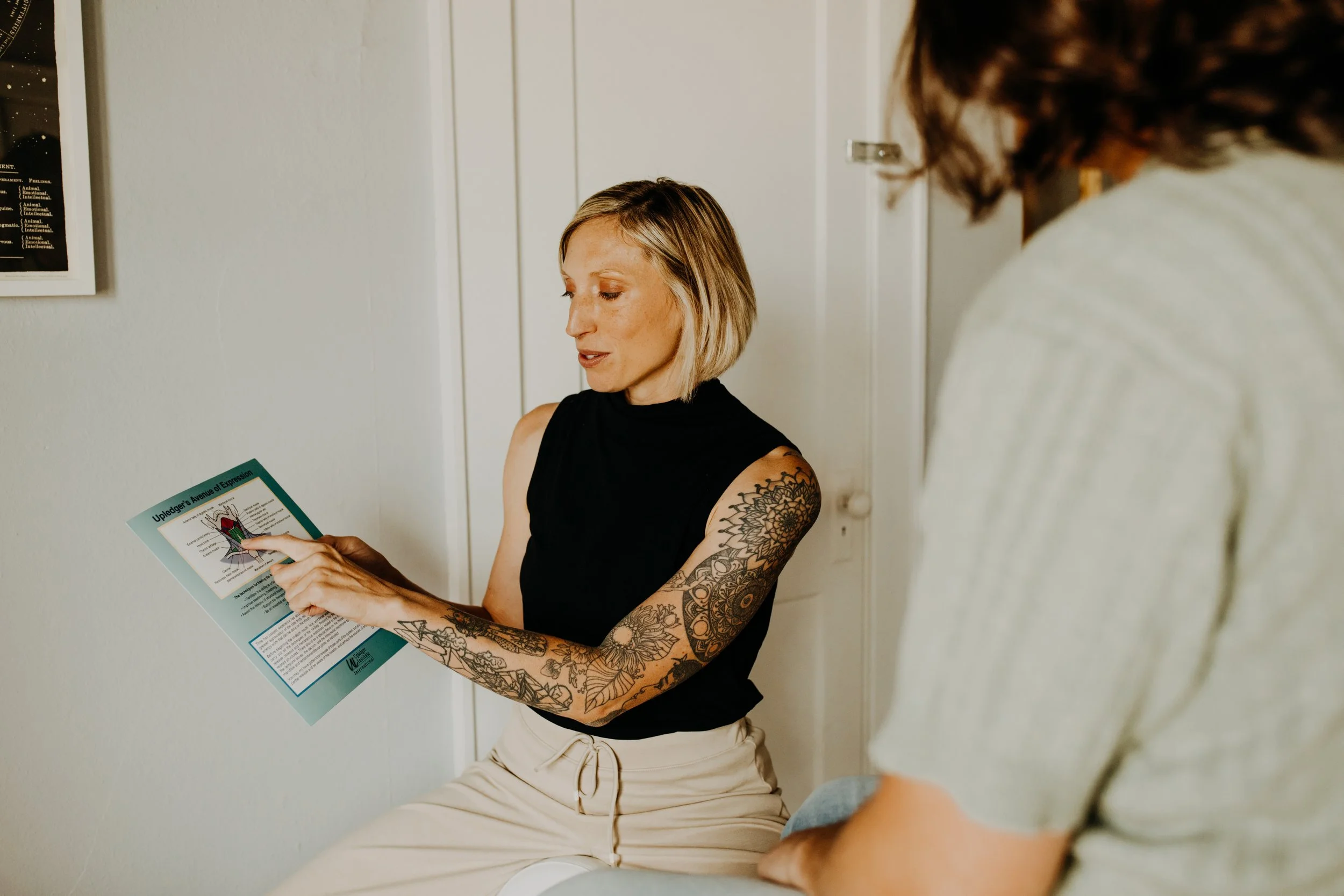 A woman with short blonde hair and tattoos on her arm is sitting on a bench, pointing to a brochure while talking to another person with long brown hair.