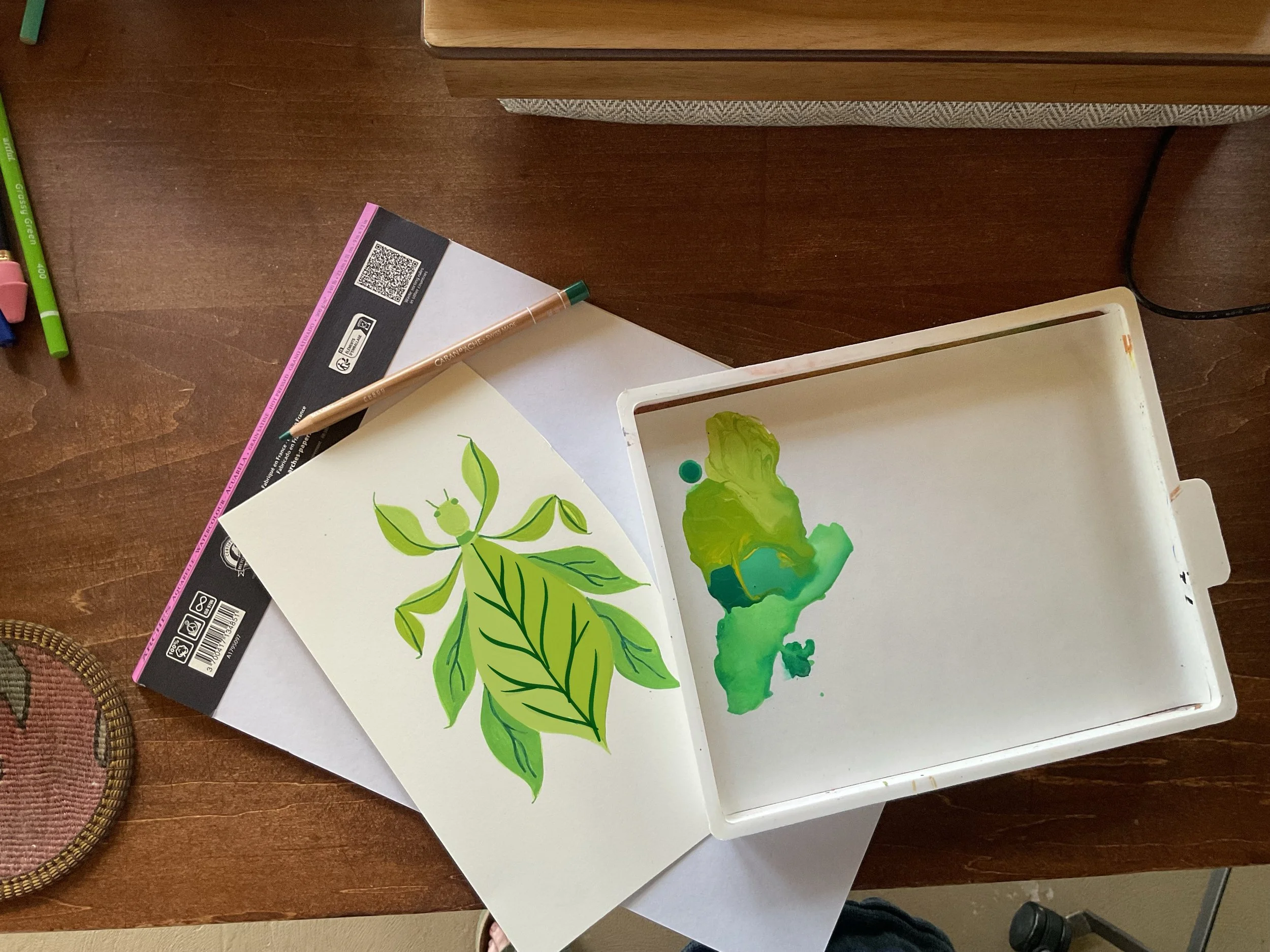 Colorful watercolor painting of green leaves and a small insect on paper, placed on a table with markers and a container nearby.