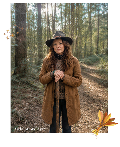 A woman with long wavy hair wearing a black hat and brown jacket stands in a forest, holding a walking stick, with tall pine trees in the background.