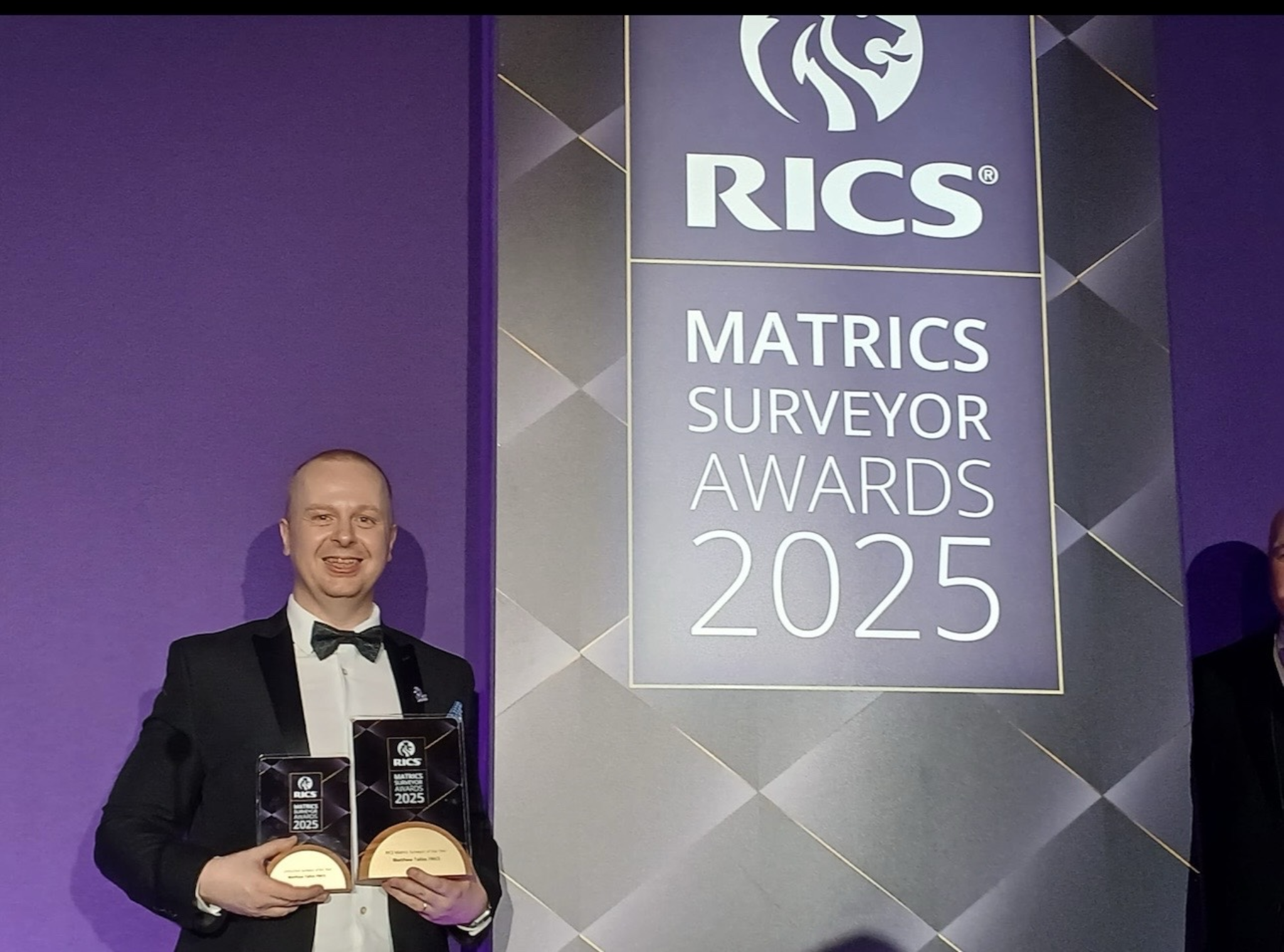 A man in a tuxedo holding awards at the RICS Matrics Surveyor Awards 2025 event, standing in front of a purple and gray backdrop with the event logo and title.