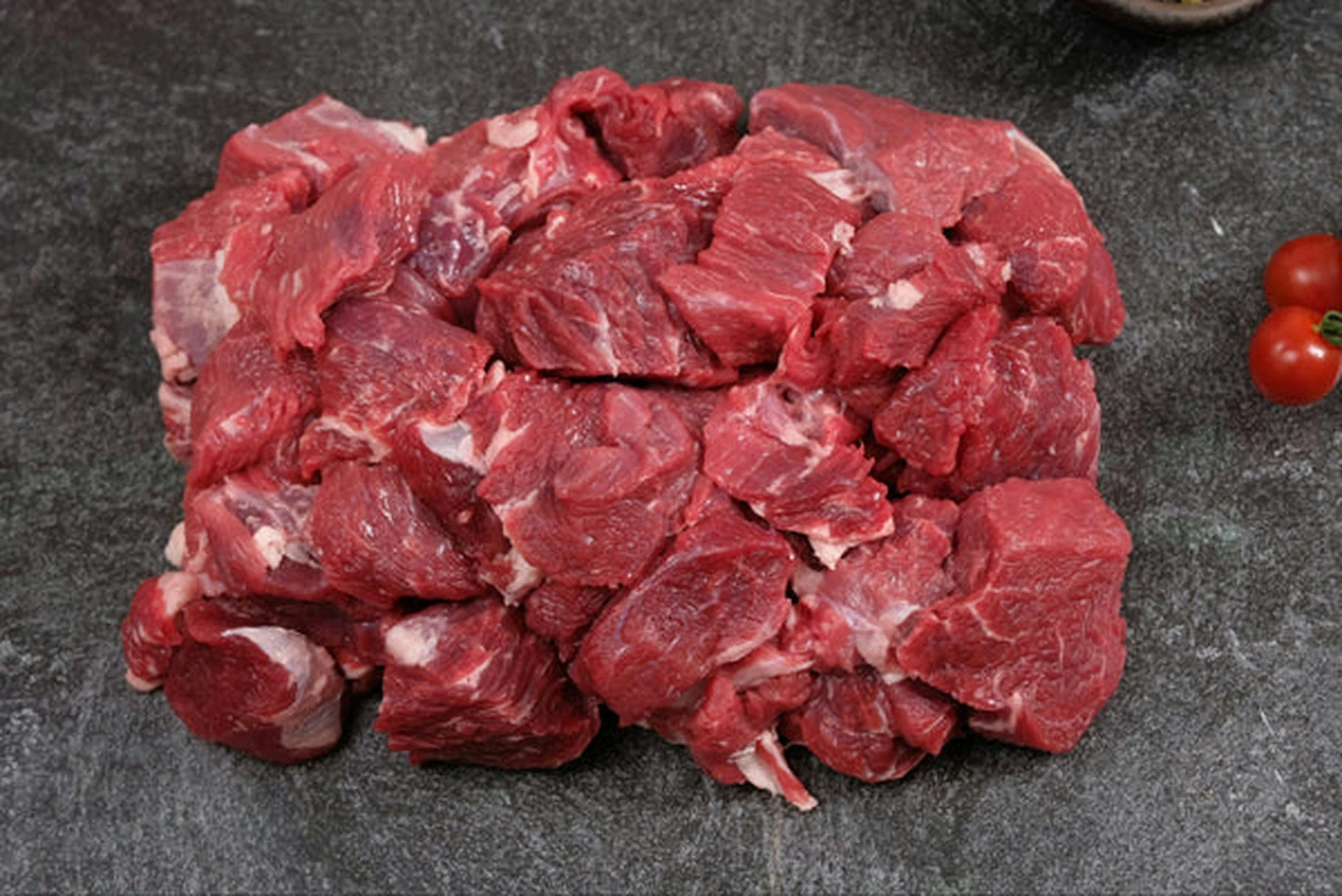 Haufen aus rohem, gewürfeltem Rindfleisch auf einer dunklen Oberfläche neben Kirschtomaten.