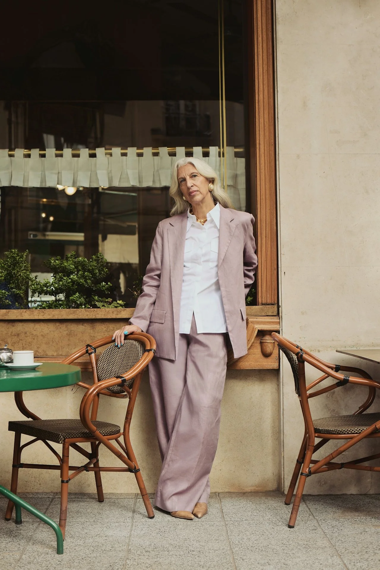 An elderly woman with long gray hair, dressed in a pale purple suit with a white shirt, standing outside a cafe or restaurant, leaning on a chair with a serious expression.