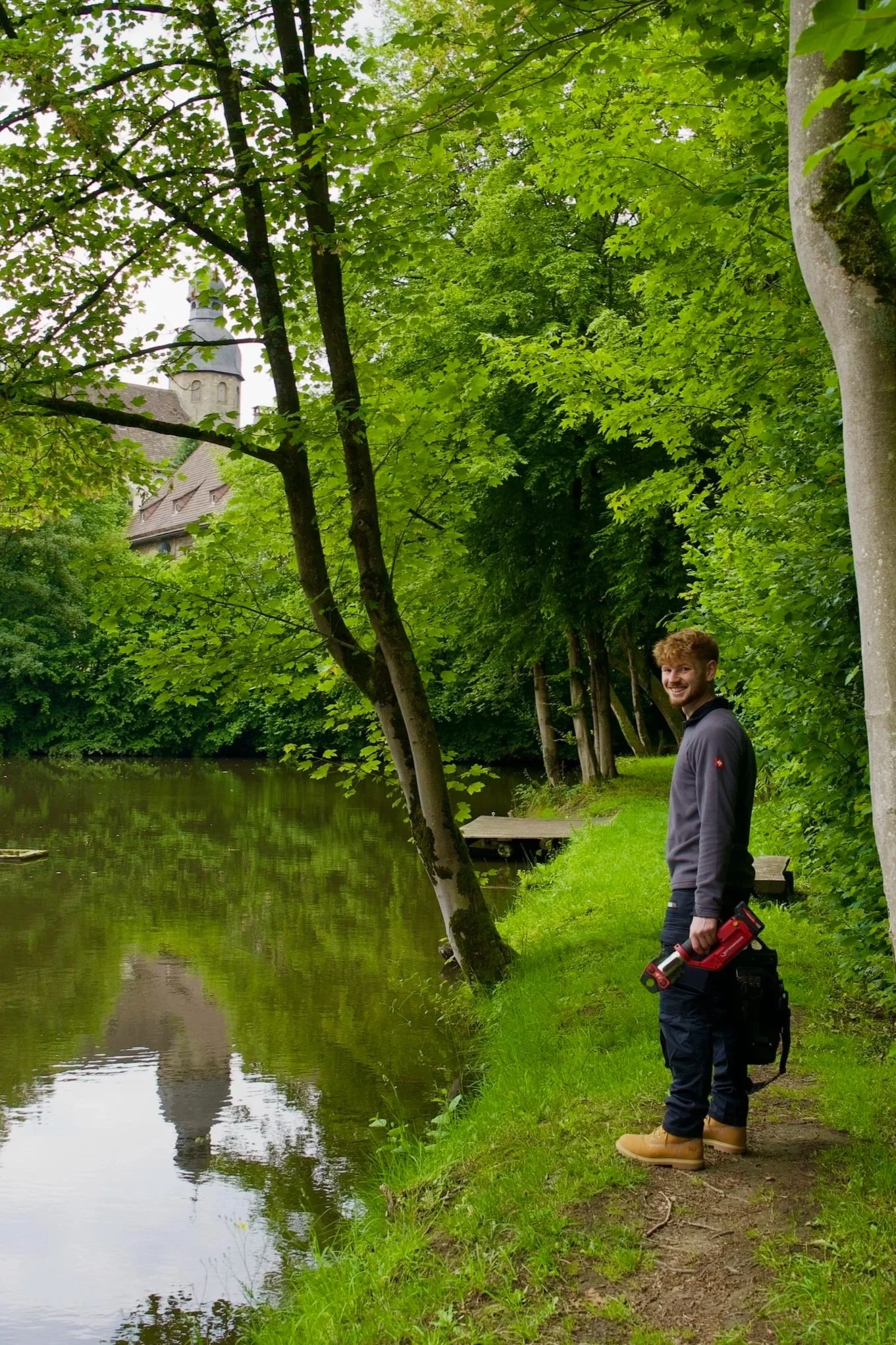 Ein Mann steht am Flussufer, trägt gelbe Gummistiefel, dunkle Hosen, eine graue Jacke, hält eine Kabelzange und eine Tasche, umgeben von grünen Bäumen und Wasser, mit einem historischen Gebäude im Hintergrund.