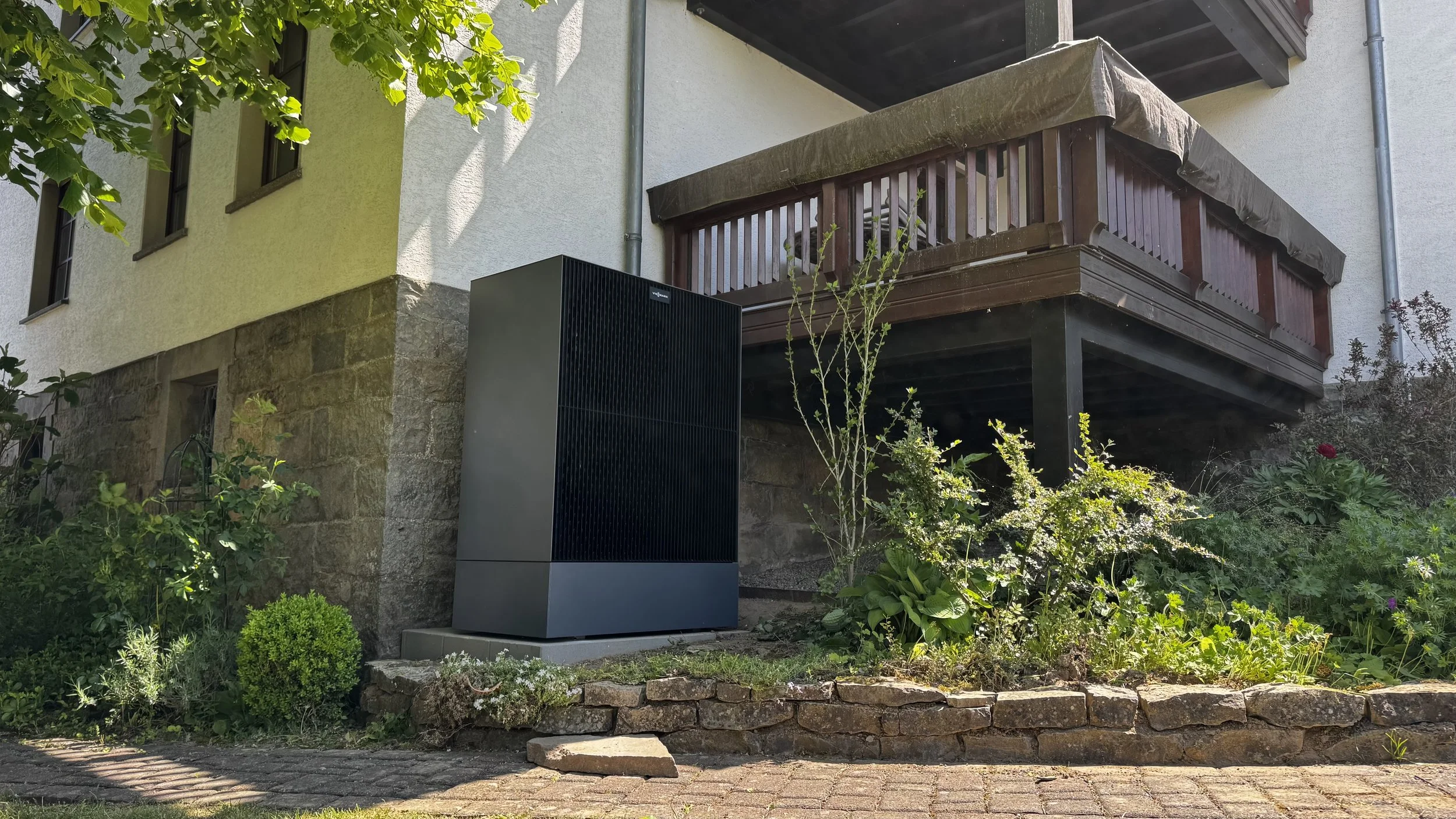 Ein Garten mit einer Hecke, Blumen und einem Haus mit Balkon. Im Vordergrund steht eine Viessmann Wärmepumpe.