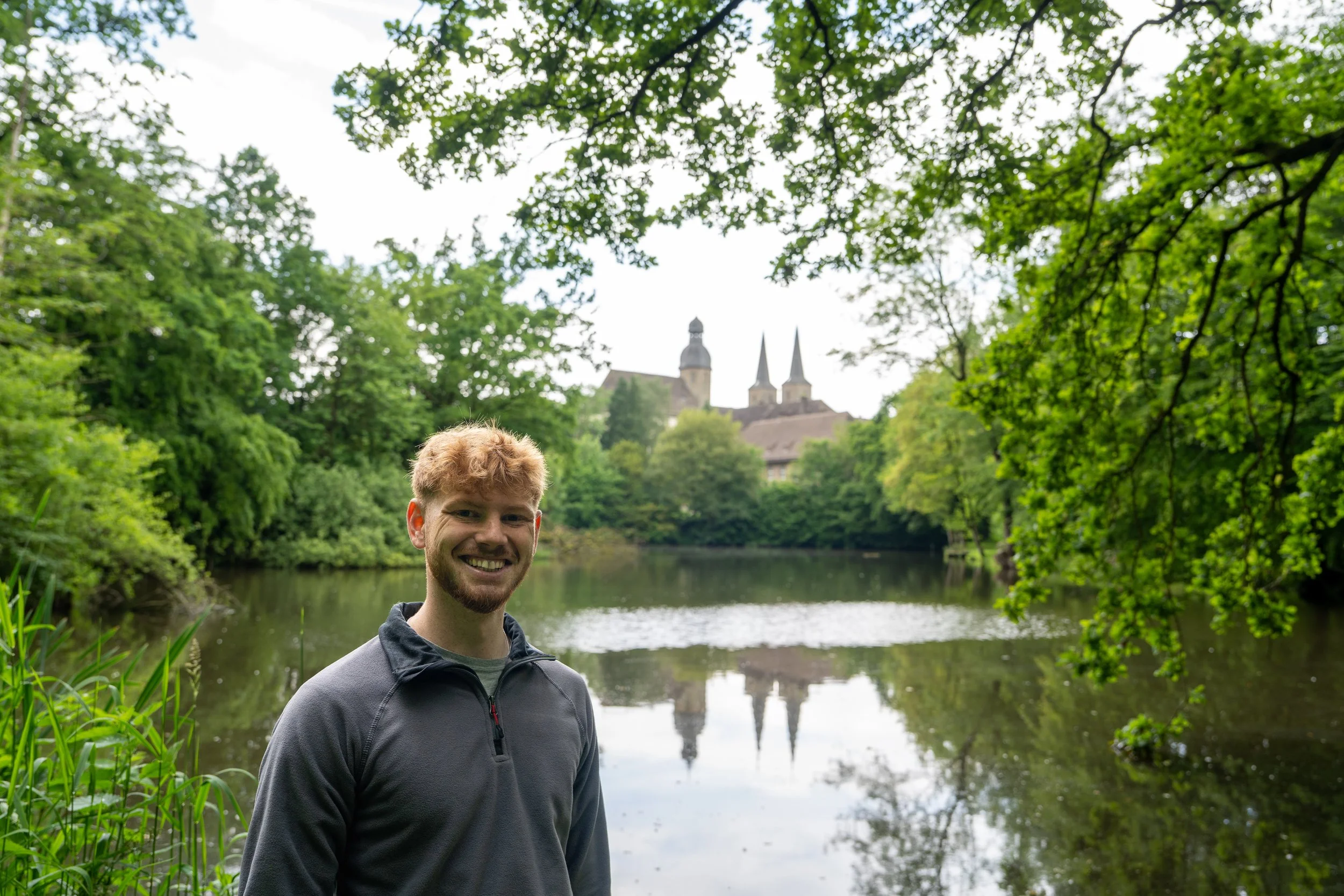 Junge Person mit dunkler Jacke, stehender Fluss im Hintergrund, grüne Bäume, Kirchen mit Türmen im Hintergrund, an einem bewölkten Tag.
