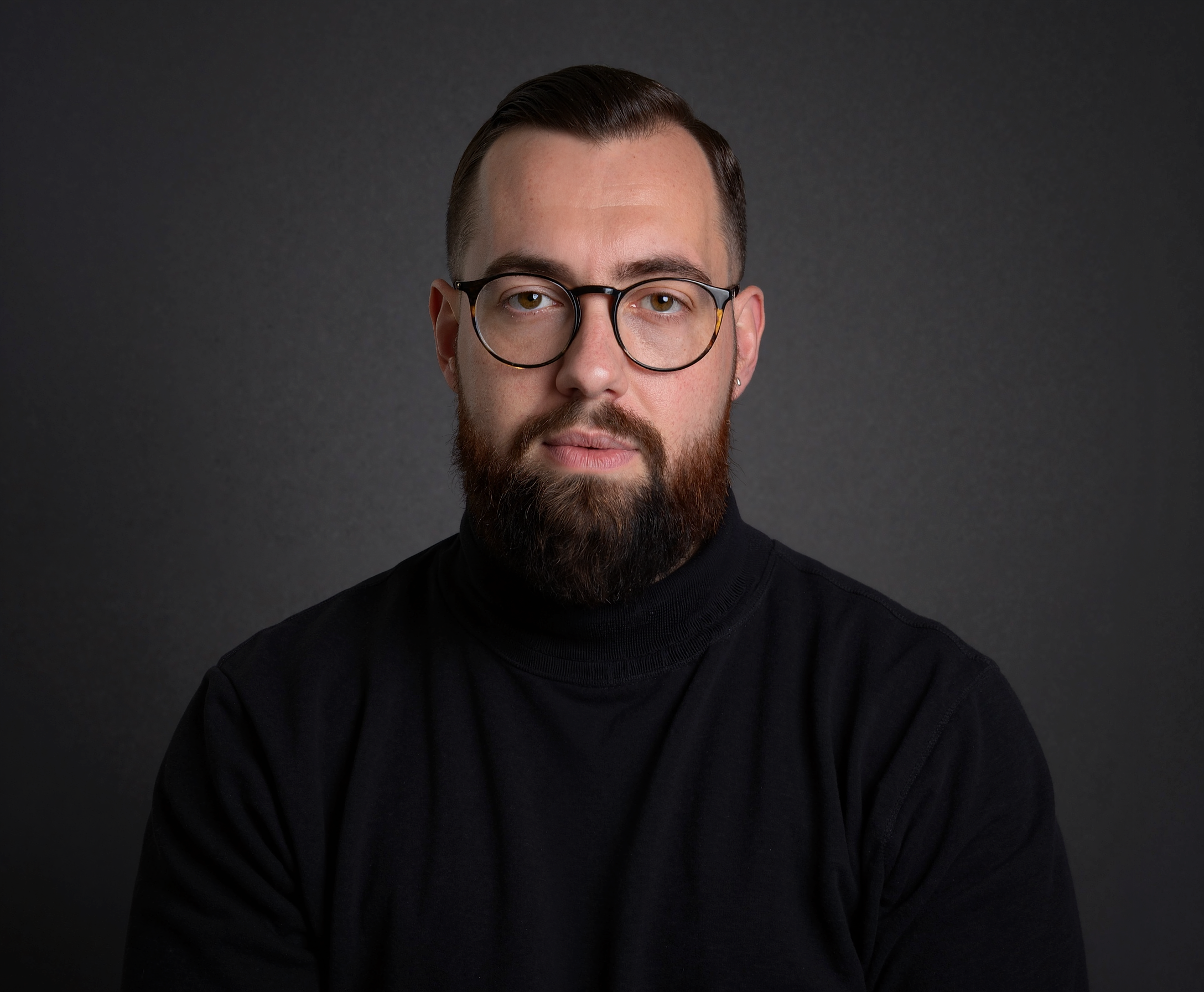 Ein Mann mit hellbraunen Haaren, Vollbart und runden Brillengläsern, trägt einen schwarzen Pullover vor dunklem Hintergrund.