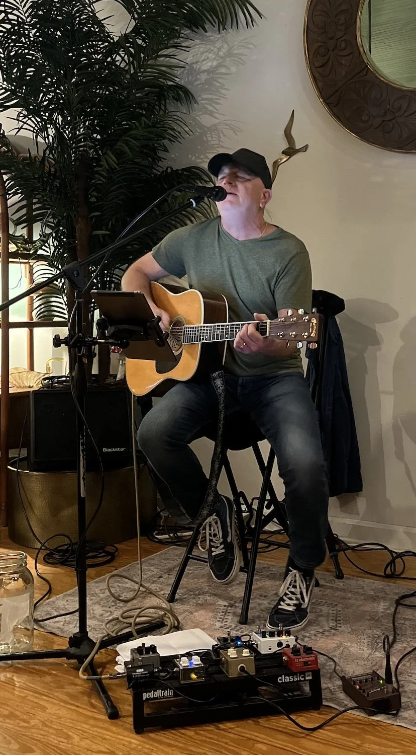 A man wearing a green t-shirt, black baseball cap, and black sneakers is sitting and playing an acoustic guitar while singing into a microphone. There is musical equipment and a large plant in the background.