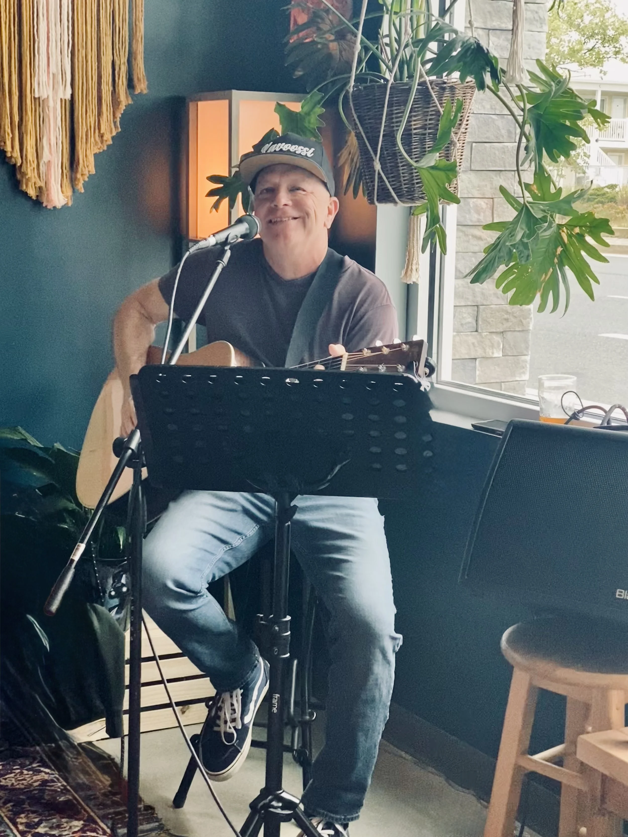 Troy Mawyer Playing Guitar, smiling, sitting on stole with big window and plant in background