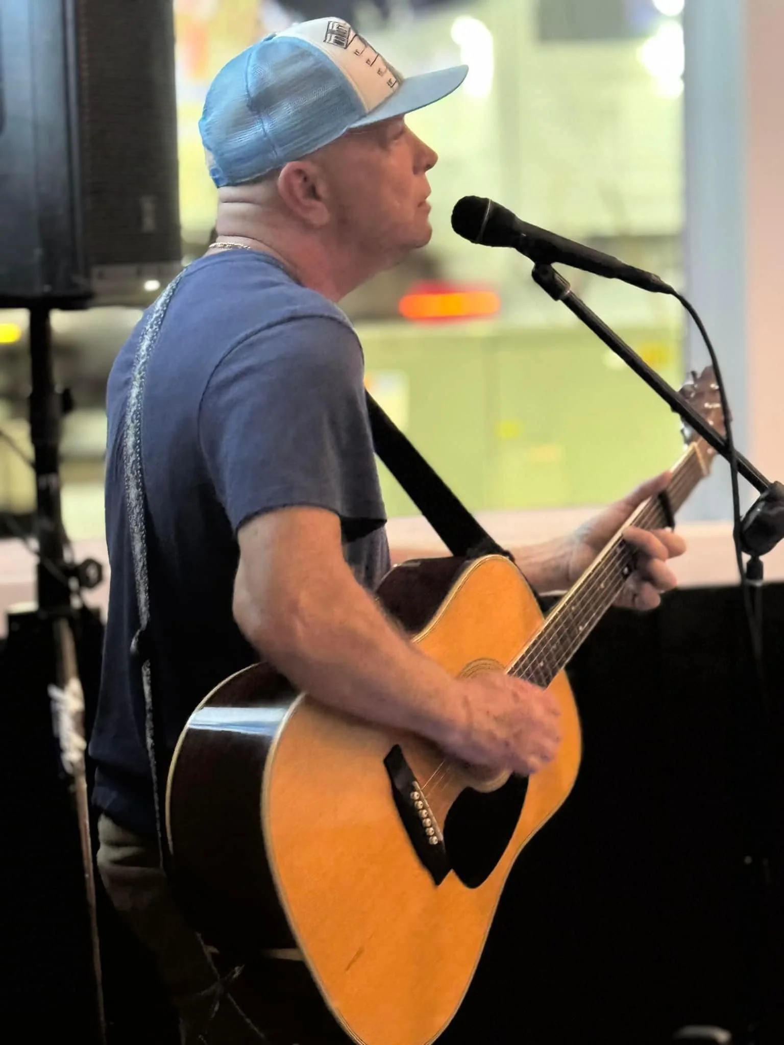 A man in a blue baseball cap and gray t-shirt playing an acoustic guitar and singing into a microphone.
