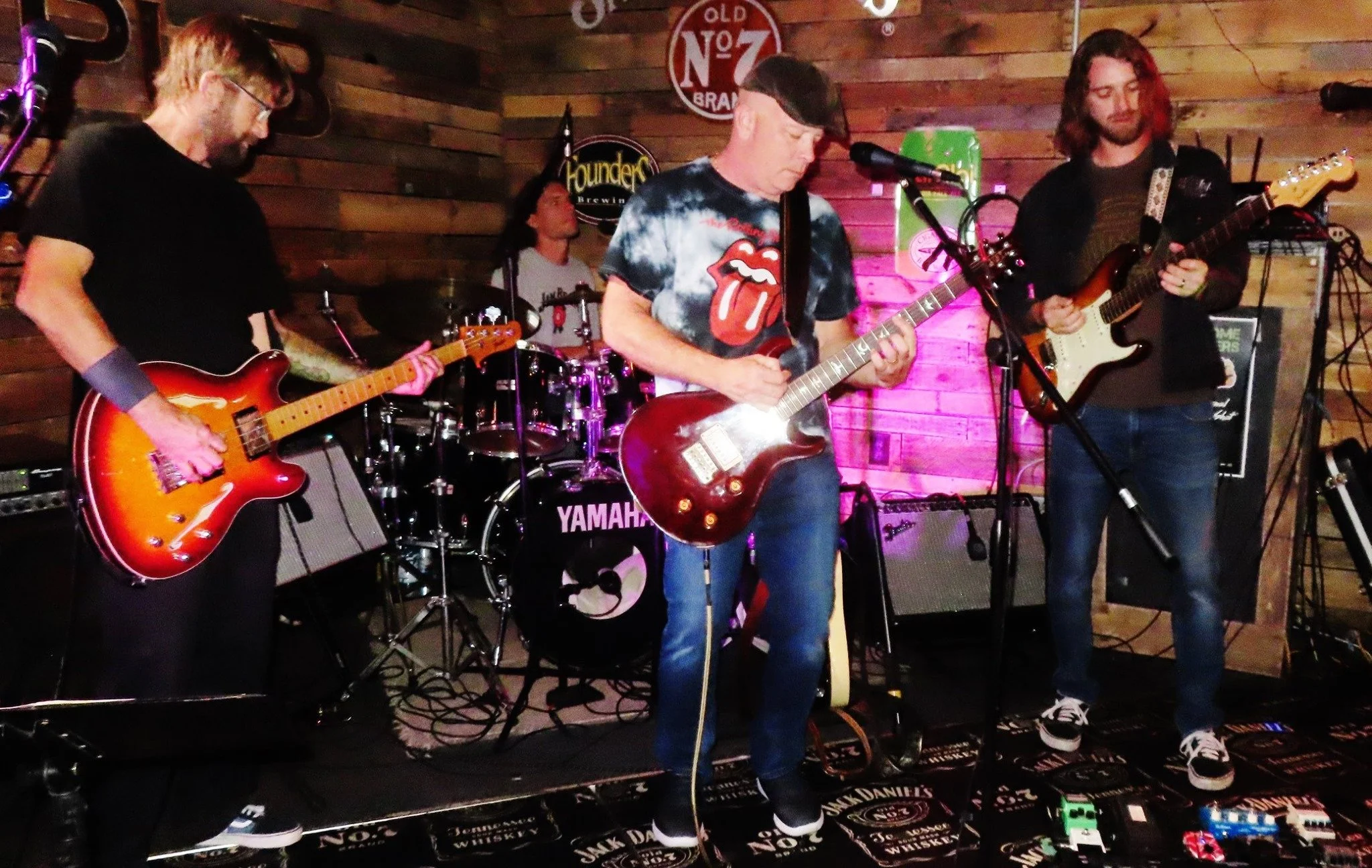 Four musicians performing on a stage with wooden wall background, with a drummer, two guitarists, and one keyboard player. They are playing electric guitars and drums, with various musical equipment and colorful lighting around them.