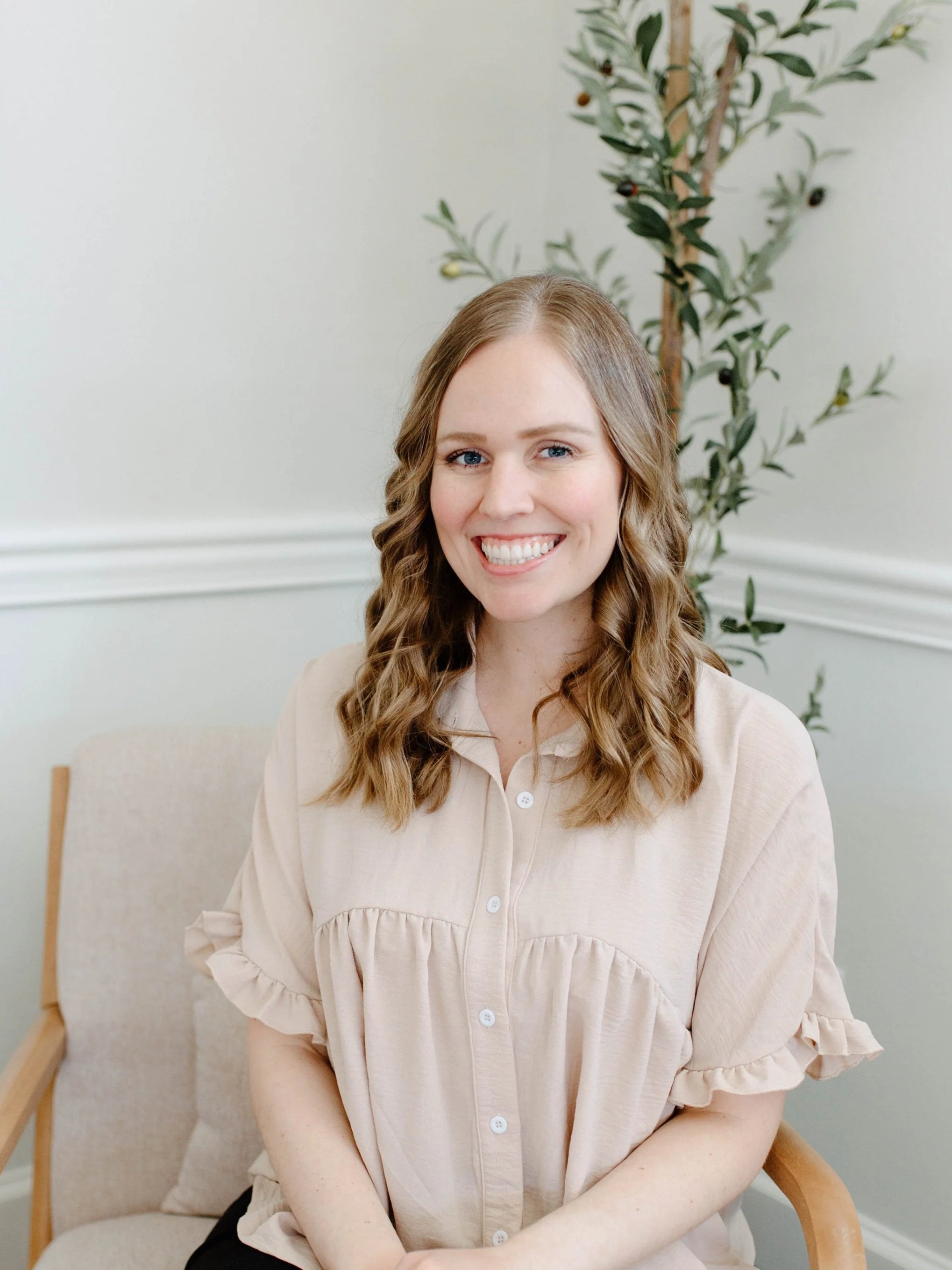 A woman with long, wavy, light brown hair smiling while sitting in a beige armchair with wooden armrests. There is a potted olive tree behind her, against a plain light-colored wall.