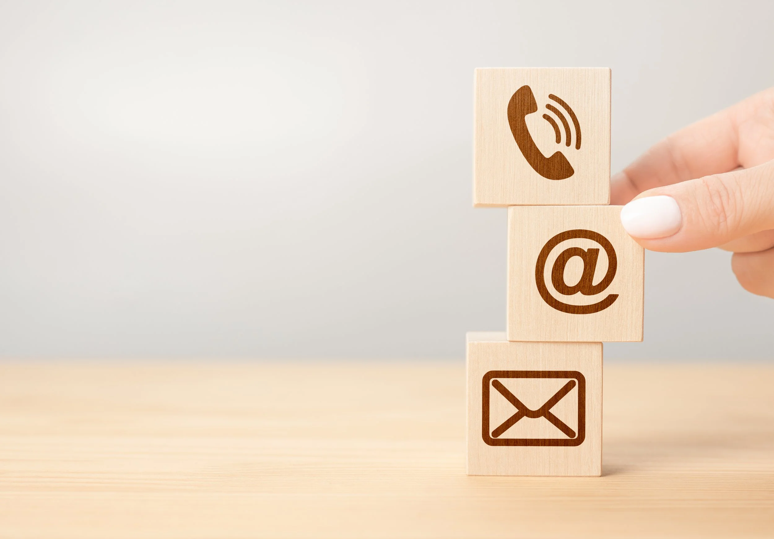 A person's hand with white nail polish holding wooden blocks with icons of a phone, email, at symbol, and envelope.